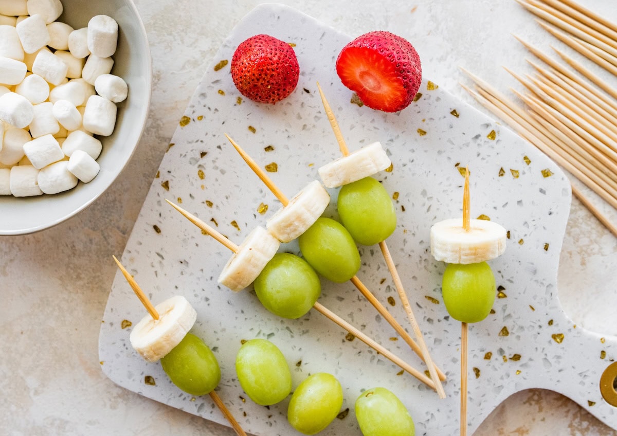Partially assembled Grinch fruit kabobs with fruit pieces threaded onto wooden skewers.