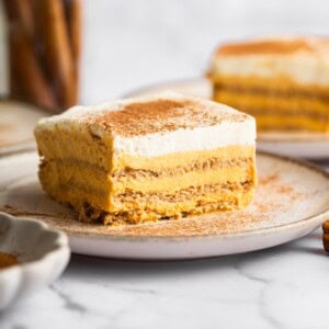A slice of pumpkin icebox cake on a plate sprinkled with cinnamon, with additional slices and a jar of cinnamon sticks in the background.