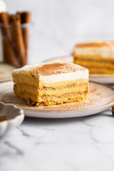 A slice of pumpkin icebox cake on a plate sprinkled with cinnamon, with additional slices and a jar of cinnamon sticks in the background.