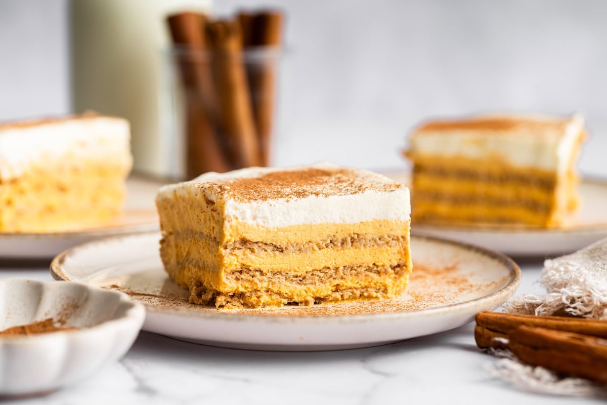 A slice of pumpkin icebox cake on a plate sprinkled with cinnamon, with additional slices and a jar of cinnamon sticks in the background.