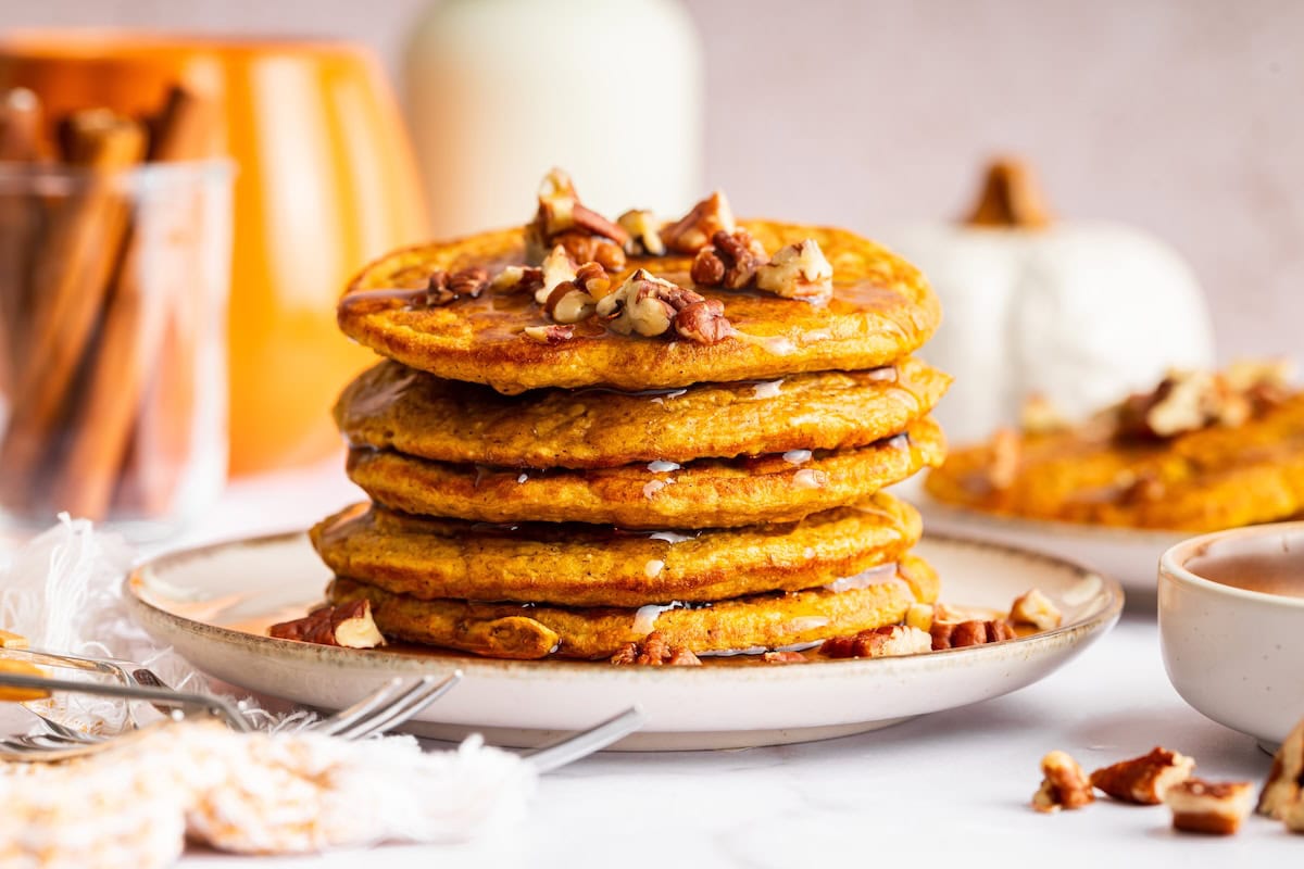 Tall stack of pumpkin protein pancakes on a plate topped with pecans and drizzled with syrup.