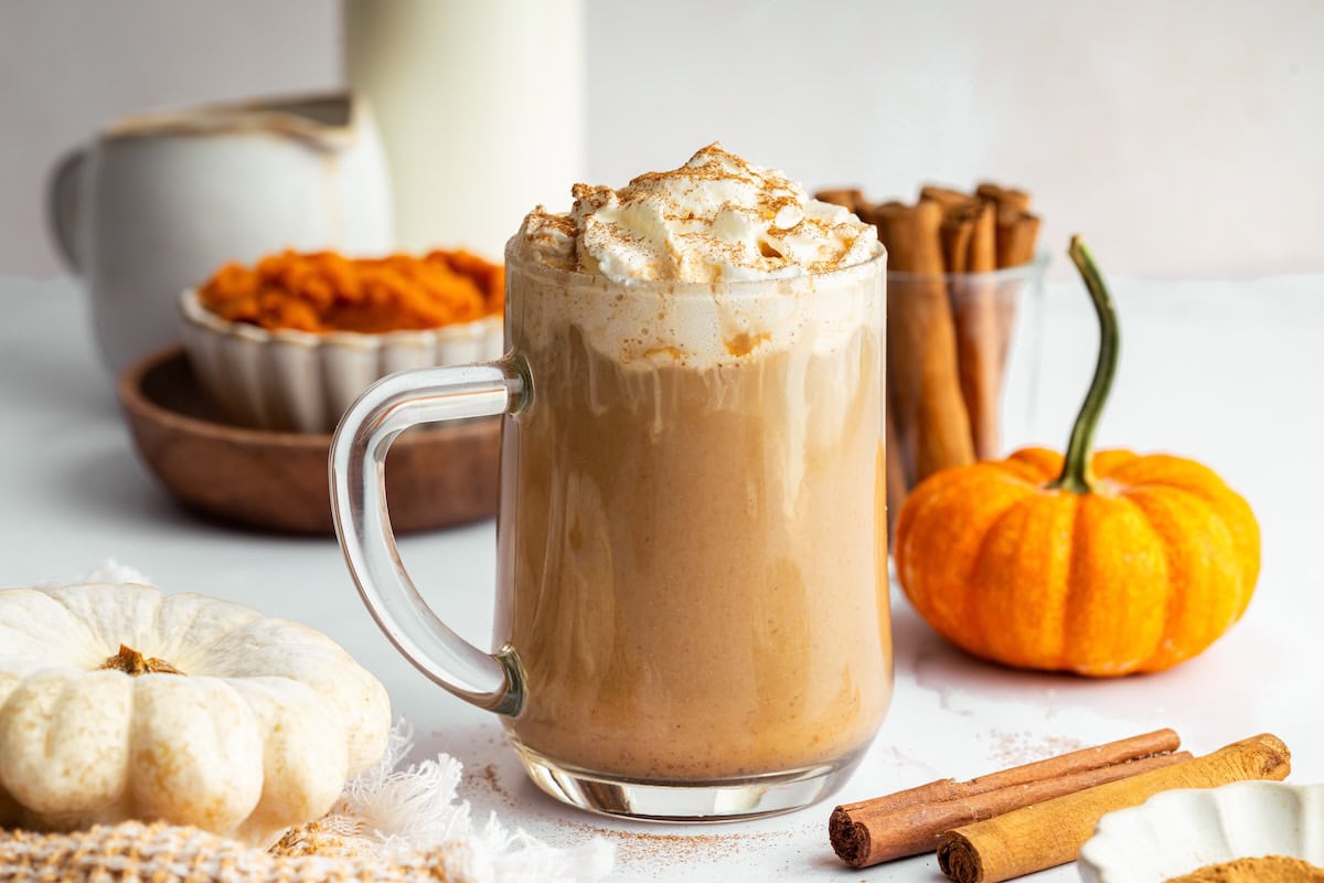 Glass mug of pumpkin spice latte topped with whipped cream and cinnamon, surrounded by pumpkins, cinnamon sticks, and a bowl of pumpkin purée.
