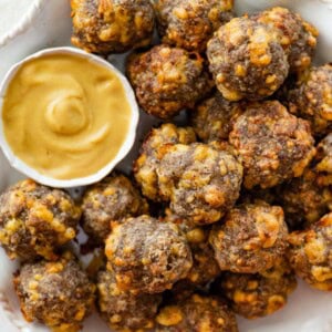 Baked sausage balls piled on a plate beside a dish of creamy mustard sauce.