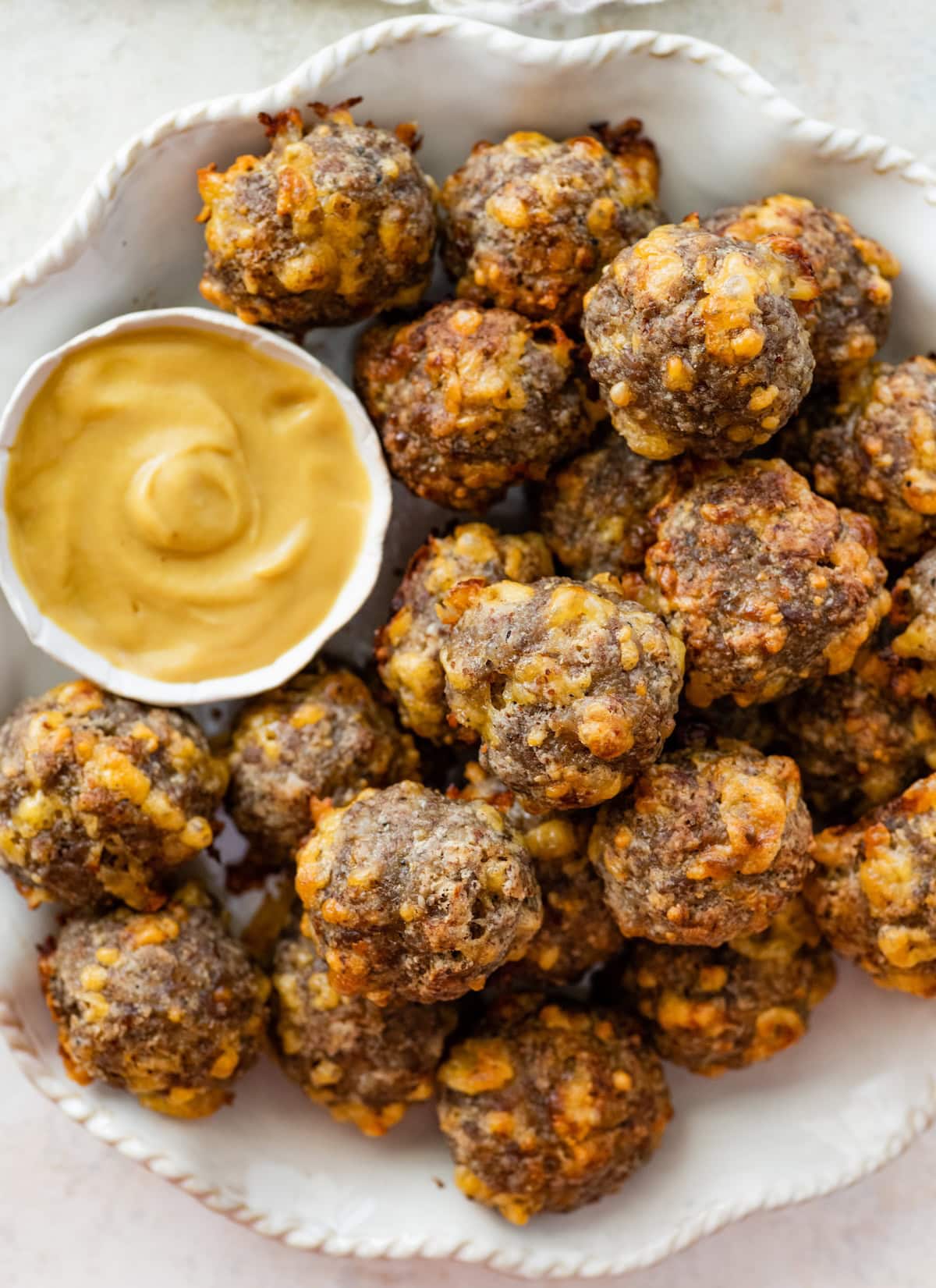 Baked sausage balls piled on a plate beside a dish of creamy mustard sauce.