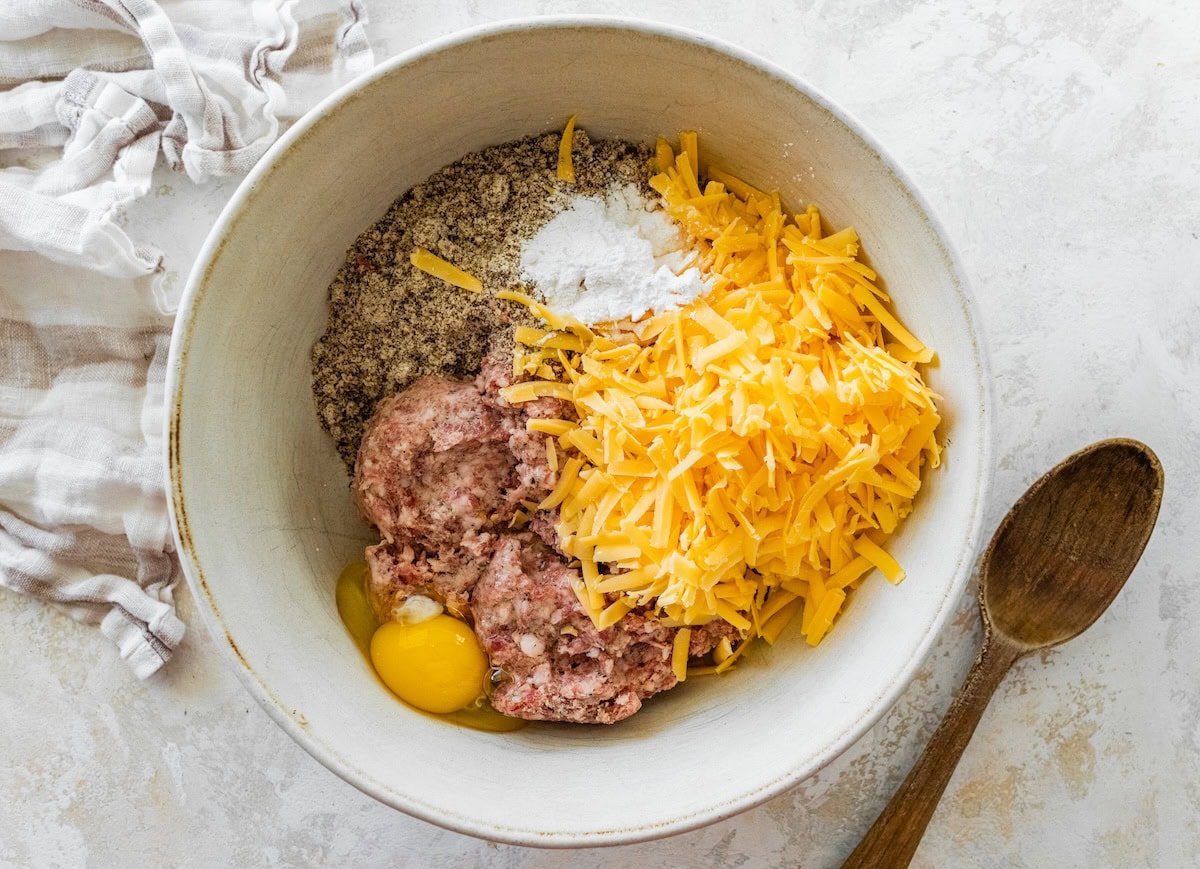 Mixing bowl filled with sausage, shredded cheddar cheese, almond flour, egg, and seasonings.