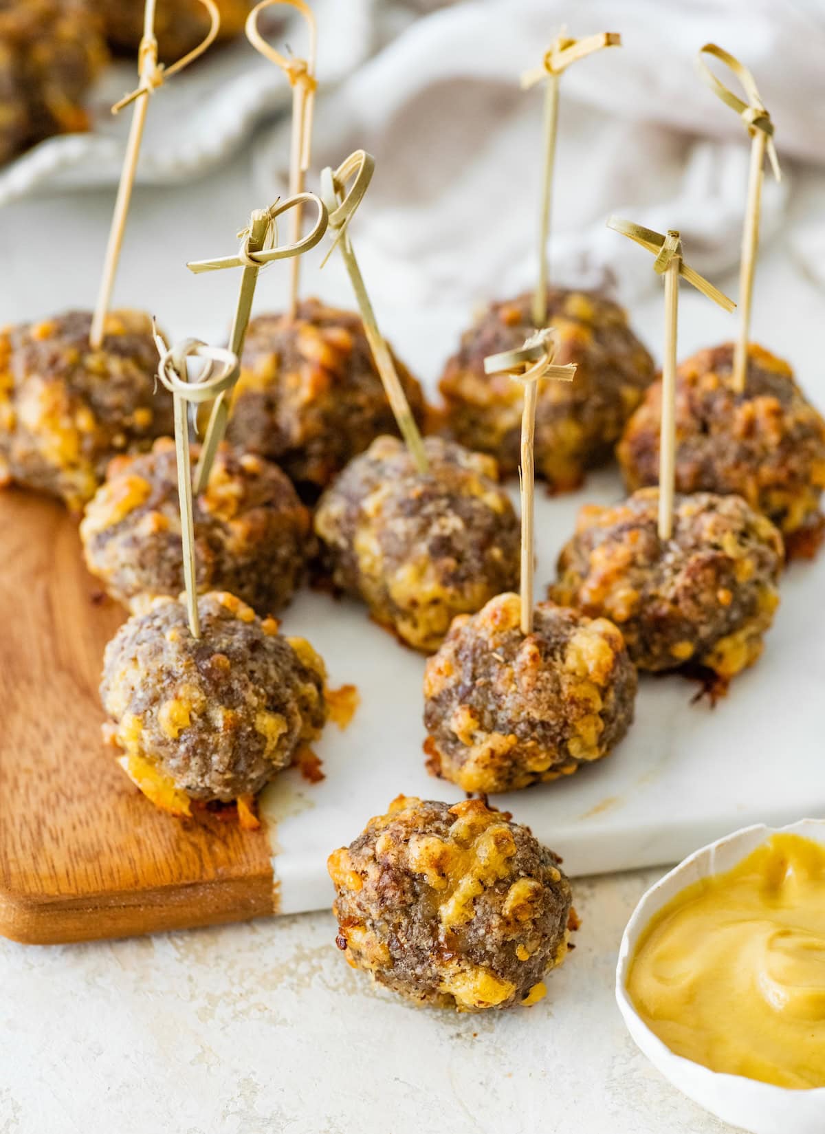 Sausage balls served on a wooden board with skewers, next to a bowl of mustard dip.