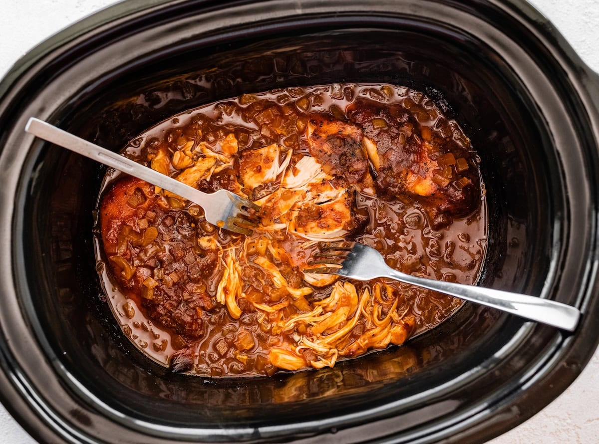 Slow cooker filled with apple BBQ chicken being shredded with two forks.