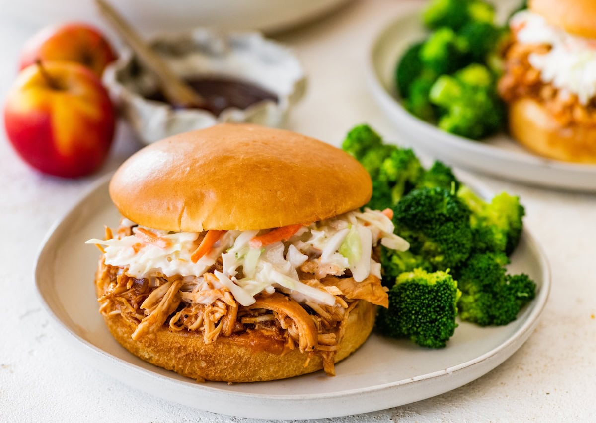 Shredded apple BBQ chicken served on a bun topped with creamy coleslaw and served with a side of broccoli.
