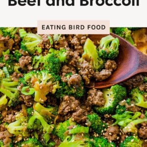 A skillet filled with cooked ground beef and broccoli florets, garnished with sesame seeds and being stirred with a wooden spoon.