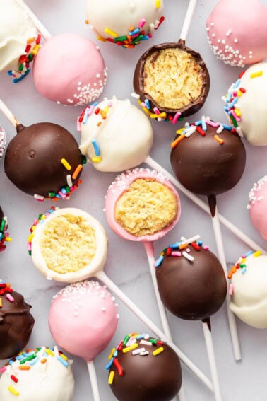 Colorful assortment of cake pops with sprinkles and white chocolate drizzle on marble background.