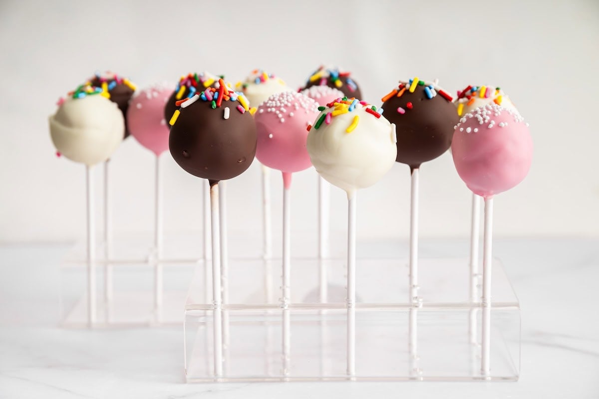 Chocolate and pink cake pops with colorful sprinkles arranged upright in a clear cake pop stand.