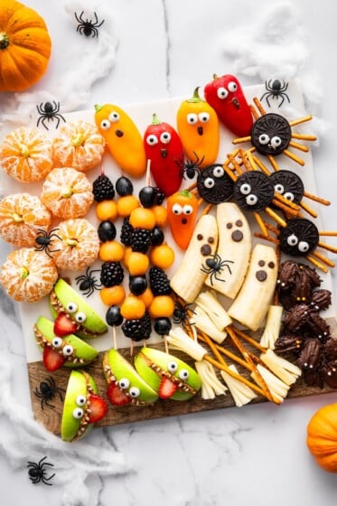 Festive Halloween snack board with banana ghosts, apple monster mouths, clementine pumpkins, Oreo spiders, veggie ghosts, and string cheese broomsticks.
