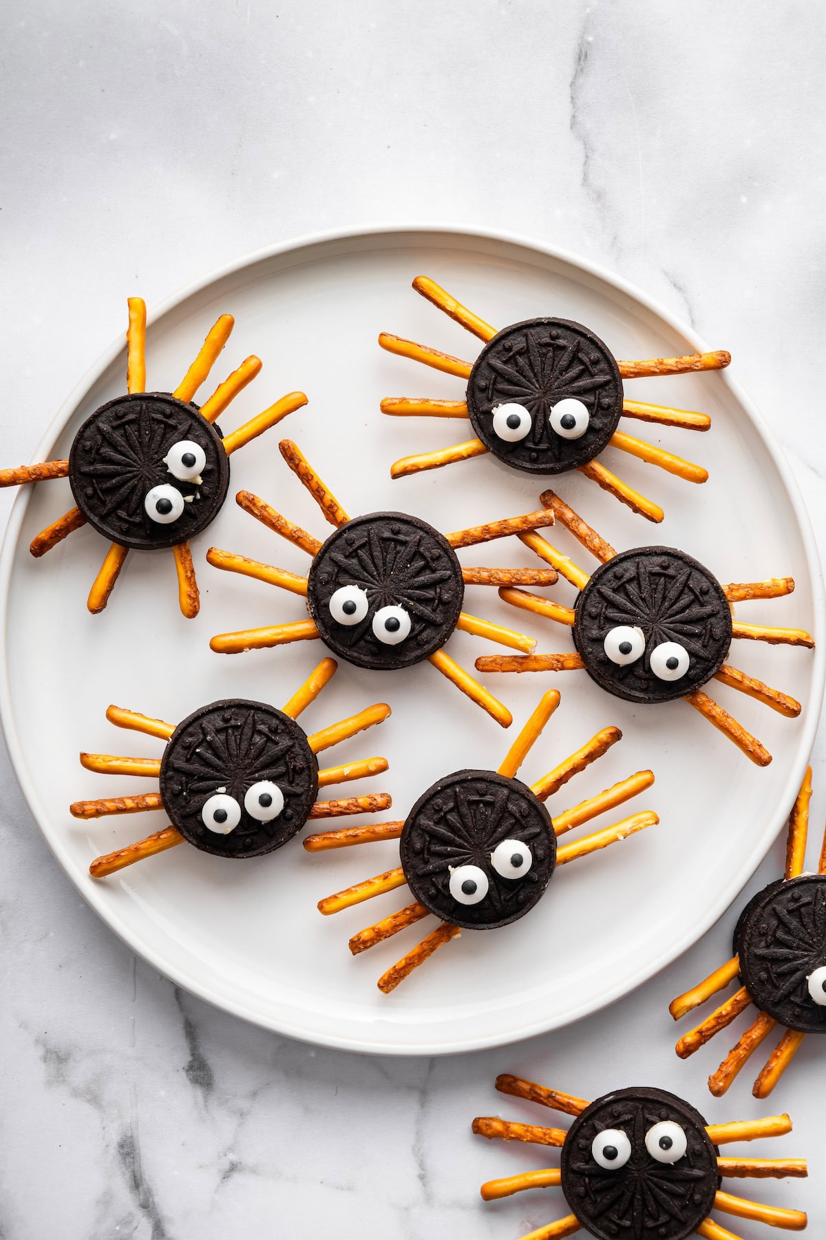Oreo spider cookies with pretzel stick legs and candy eyes arranged on a white plate.