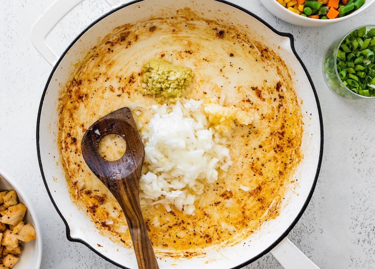 Diced onion, garlic, and ginger sautéing in a skillet with a wooden spoon.