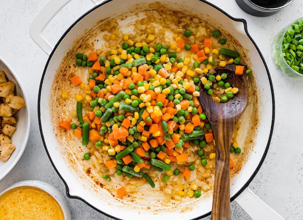 Frozen mixed vegetables added to a saucepan with sauteed onions, garlic and ginger with a wooden spoon.