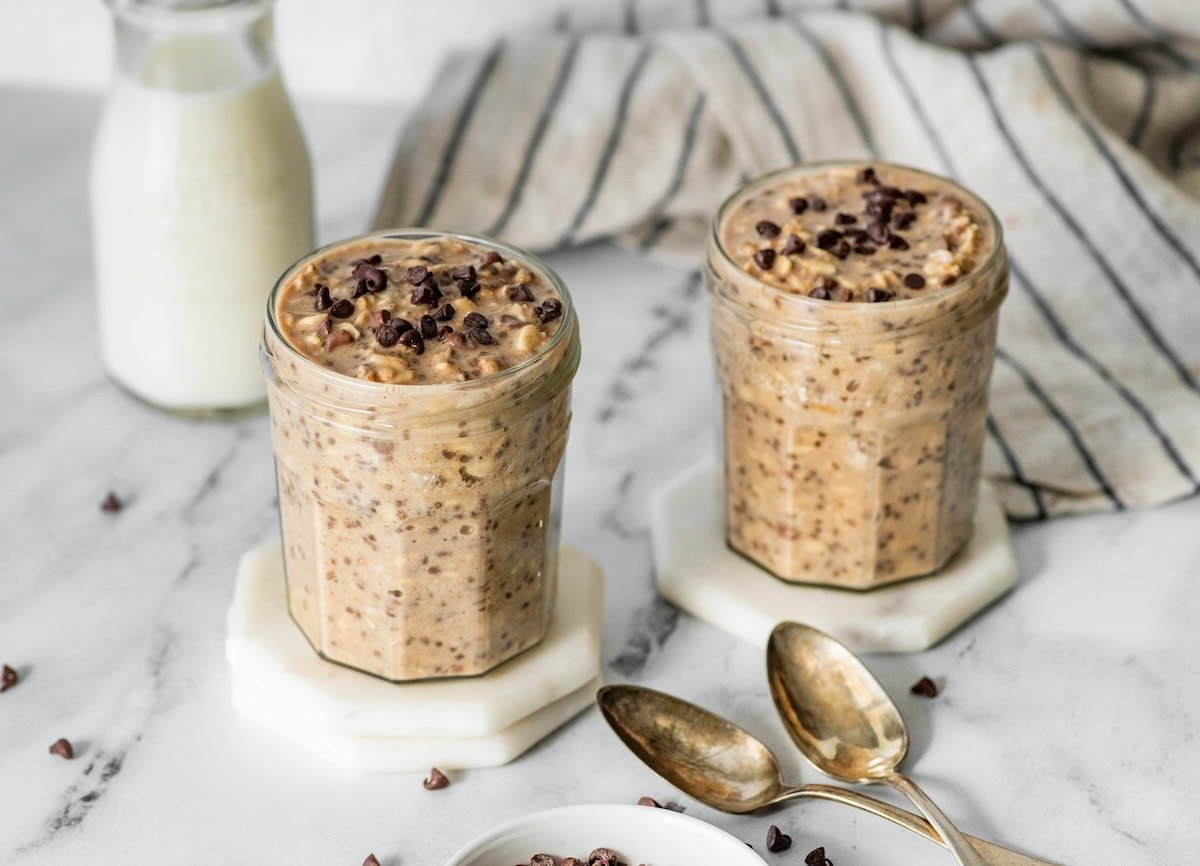 Two jars of cookie dough overnight oats with chocolate chips and spoons nearby.