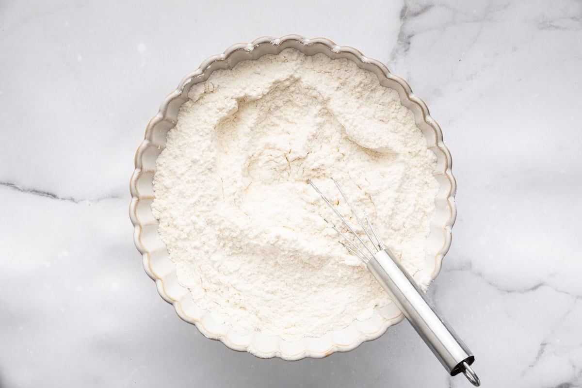 A shallow bowl filled with the dry ingredients for sugar cookies—flour, baking powder, and salt—being whisked together with a metal whisk.