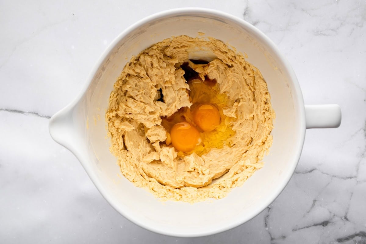 A mixing bowl filled with creamed butter and sugar, with two cracked eggs and vanilla sitting on top, ready to be blended in.