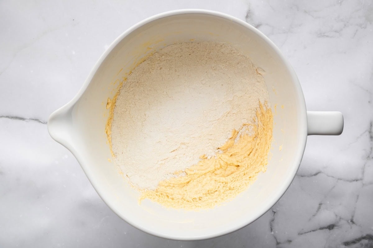 A large white mixing bowl holding partially mixed sugar cookie dough with a mound of flour sitting on top.