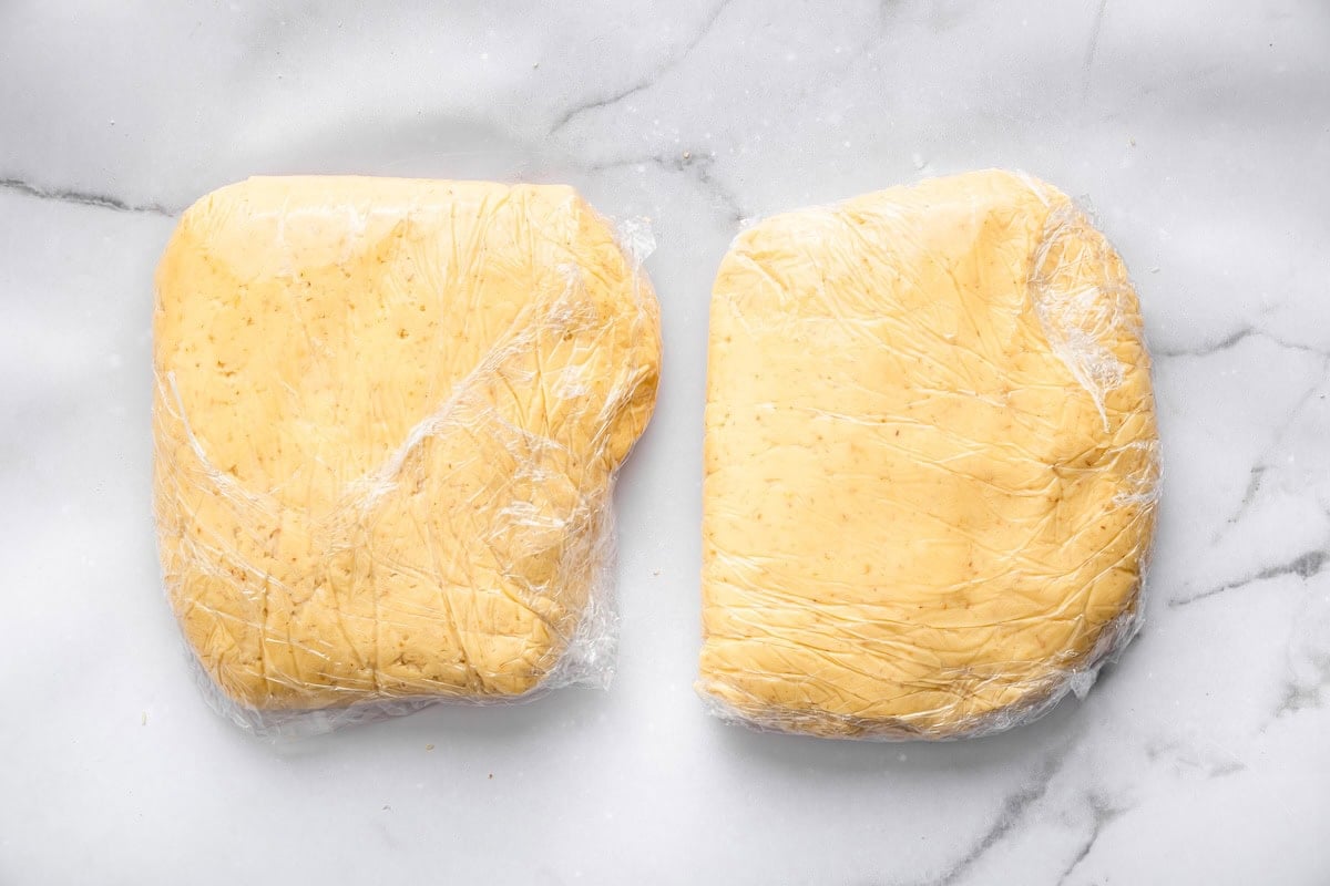 Two flattened discs of sugar cookie dough wrapped tightly in plastic wrap resting on a white marble surface. The dough is smooth, pale yellow, and ready to be chilled before rolling.