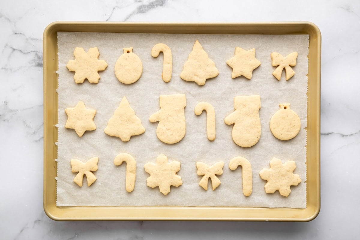 A parchment-lined baking sheet filled with unbaked sugar cookie cutouts shaped like stars, snowflakes, trees, mittens, candy canes, bows, and ornaments, arranged neatly before baking.