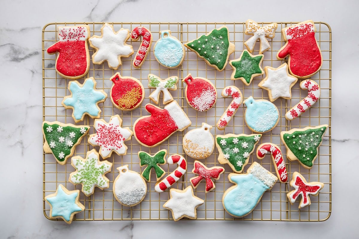 A cooling rack filled with iced holiday sugar cookies in bright colors, including trees, mittens, snowflakes, ornaments, bows, and candy canes.