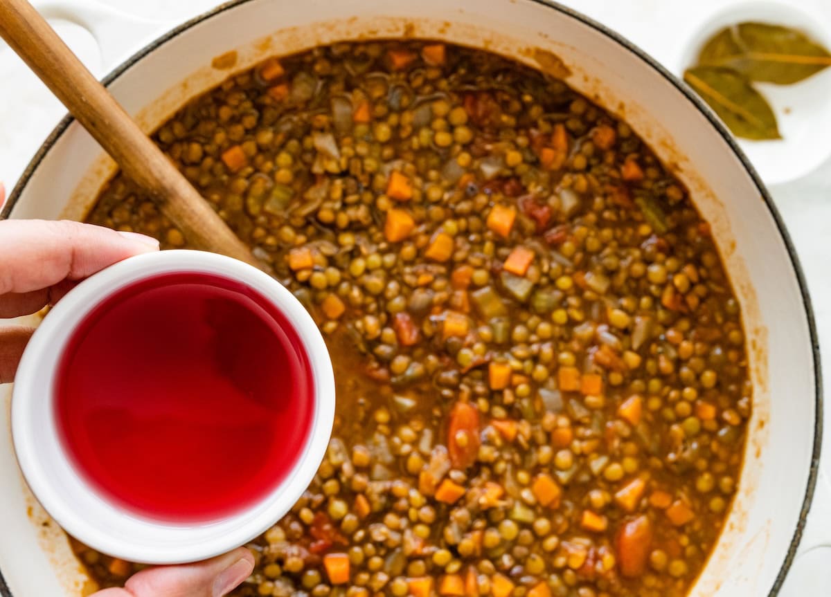A hand holds a small container of red wine vinegar over a large pot of lentil soup.