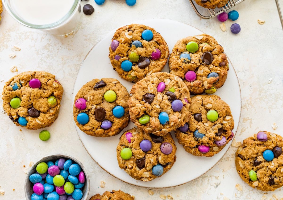 A full plate of monster cookies surrounded by scattered candies, a cooling rack, and two glasses of milk on the table.