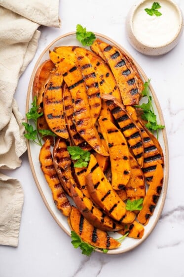 Grilled sweet potatoes on a white platter garnished with fresh parsley and served alongside a garlic aioli dip.