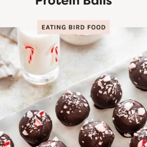 Chocolate peppermint protein balls coated in dark chocolate and topped with crushed candy cane, arranged on a tray next to a glass of milk with candy cane decorations.