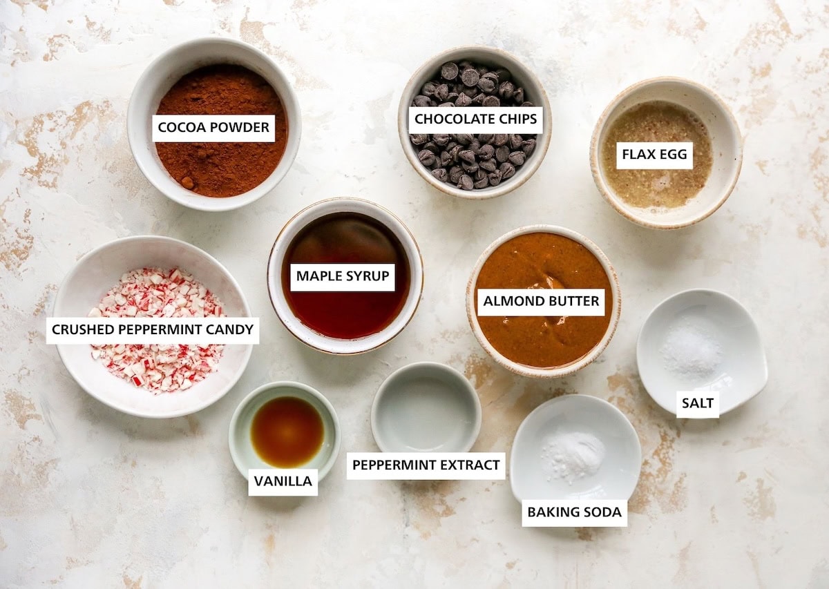 Ingredients measured out to make chocolate peppermint cookies: flax egg, salt, baking soda, almond butter, chocolate chips, peppermint extract, maple syrup, vanilla, cocoa powder and crushed peppermint candy.