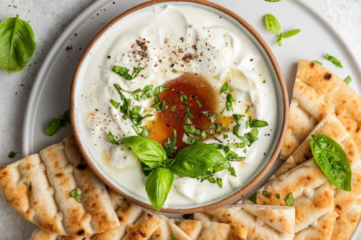 Whipped feta dip in a bowl, on a plate, near many slices of pita bread.
