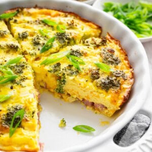 Crustless quiche with broccoli, cheese, onion, and a green onion garnish in a round baking dish with a slice removed.