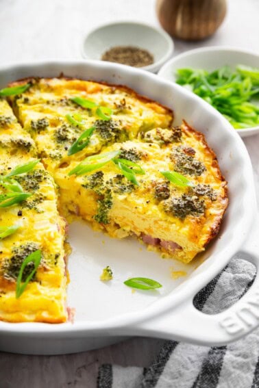 Crustless quiche with broccoli, cheese, onion, and a green onion garnish in a round baking dish with a slice removed.