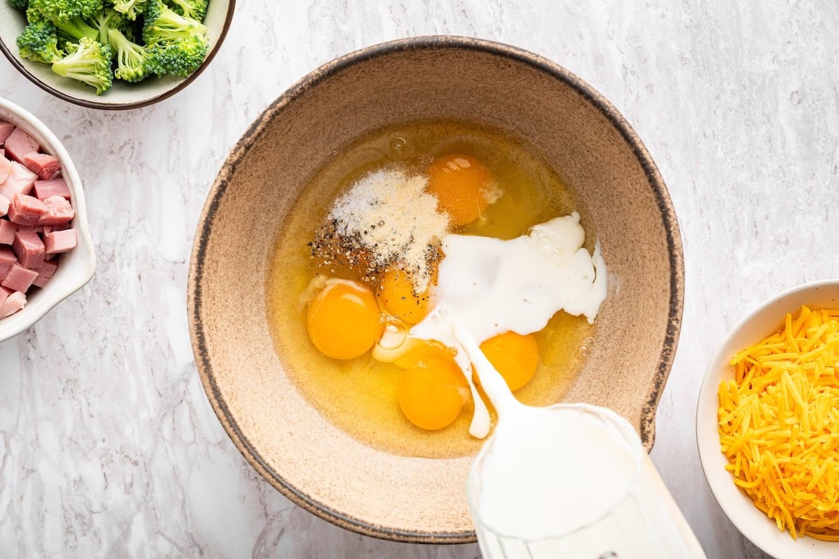 Milk being poured into a large mixing bowl containing eggs and seasonings.