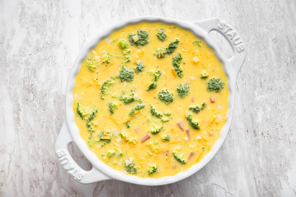A crustless quiche with broccoli, ham, onion, and cheese in a round baking dish before being baked.