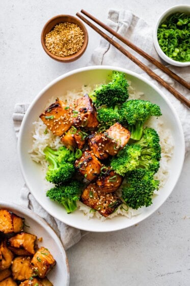 Teryaki salmon bites served on a plate with white rice and broccoli topped with sesame seeds and green onions.