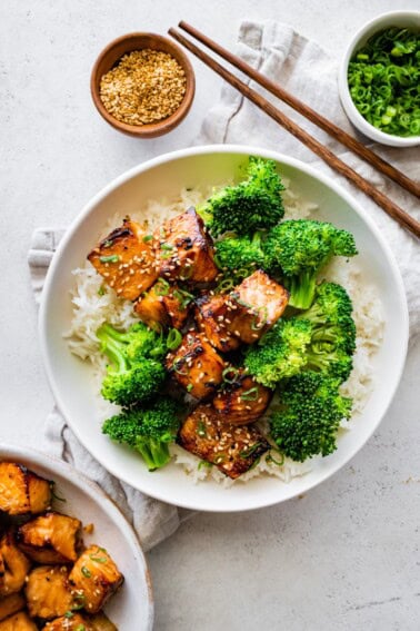 Teryaki salmon bites served on a plate with white rice and broccoli topped with sesame seeds and green onions.