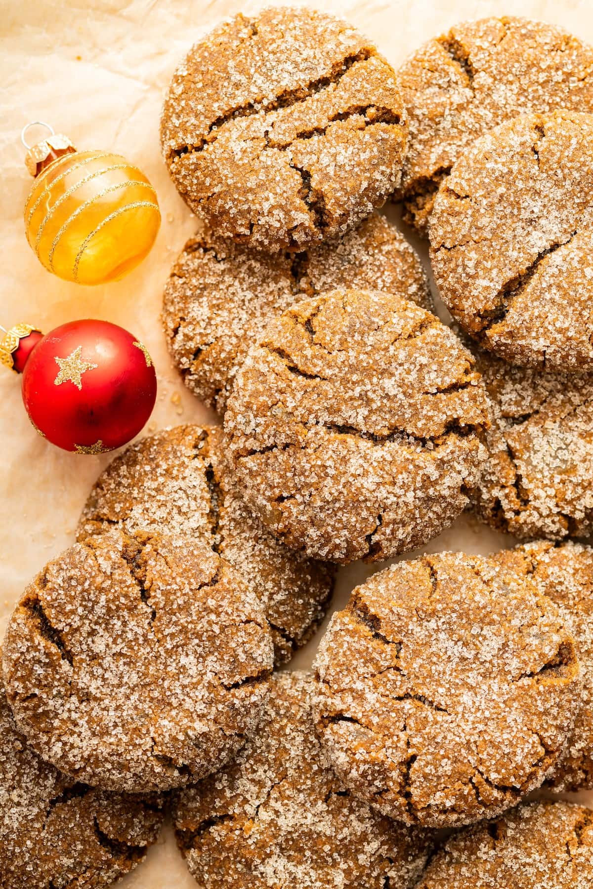 Multiple gingersnap cookies stacked over one another on parchment paper with two small ornaments nearby.