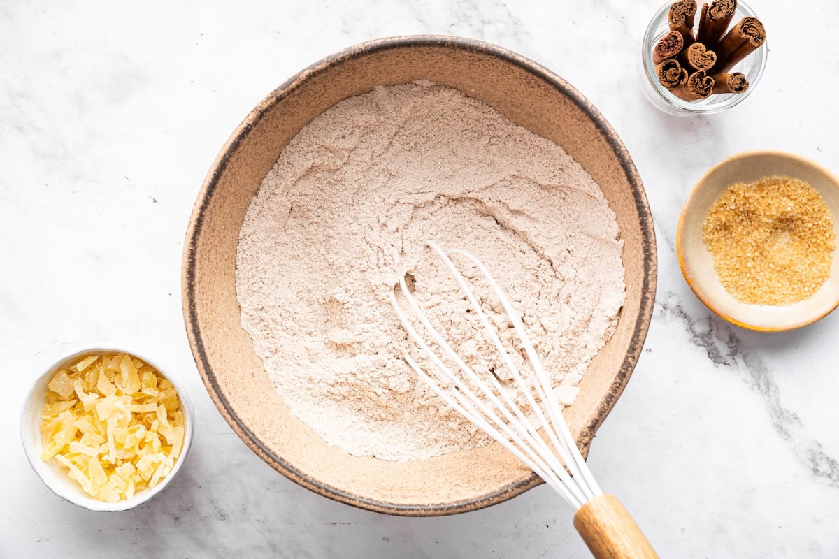 A medium mixing bowl of dry ingredients for gingersnap cookies with a whisk and a small bowl of crystalized ginger, a small bowl of a flax egg, and a small container of cinnamon sticks to the side.