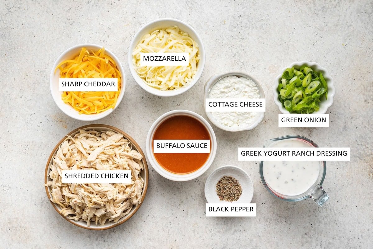 Ingredients measured out to make Healthy Buffalo Chicken Dip: shredded chicken, cheddar, buffalo sauce, mozzarella, black pepper, cottage cheese, Greek yogurt ranch dressing and green onion.