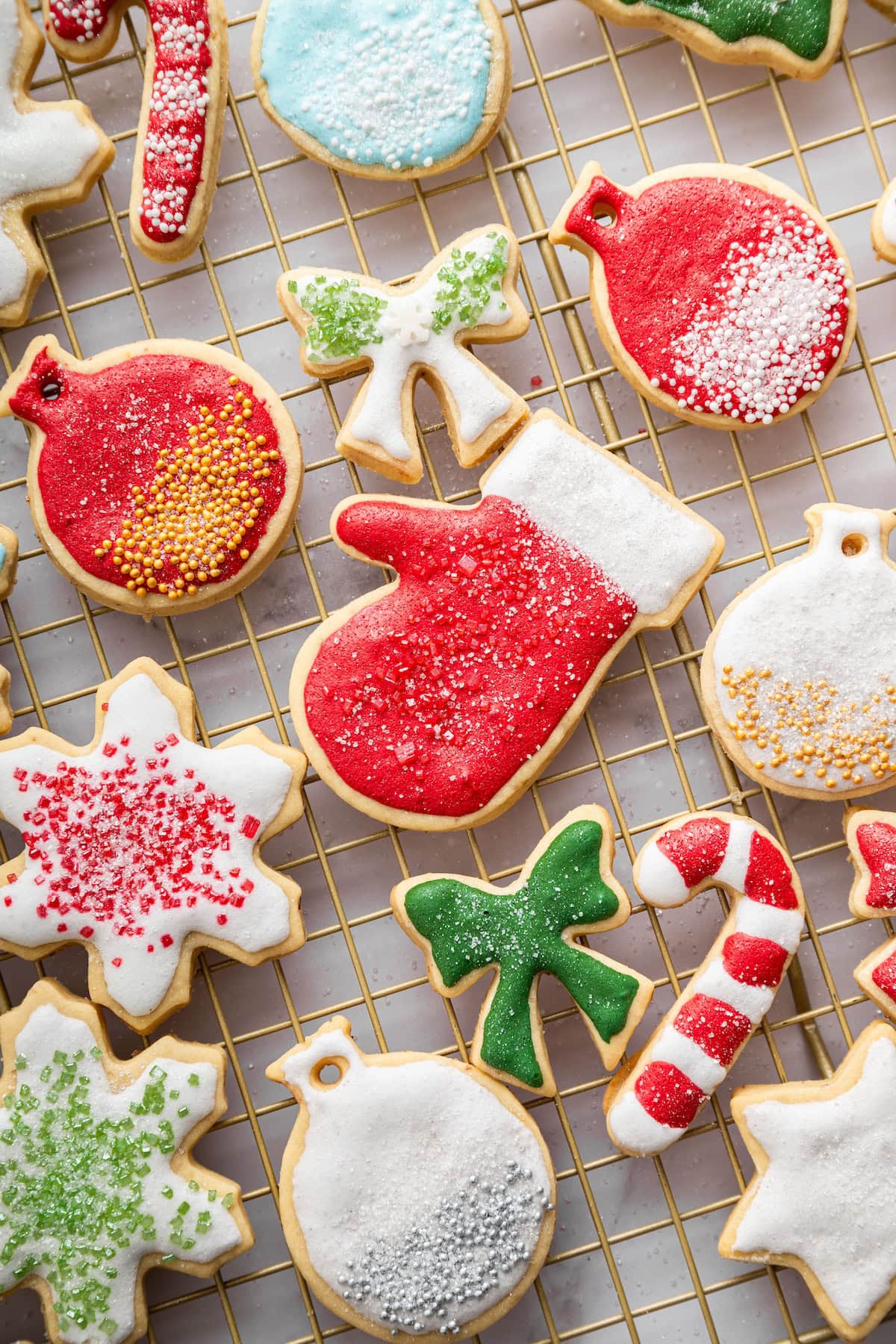 Decorated holiday sugar cookies in red, green, blue, and white icing spread across a cooling rack, showing shapes like mittens, bows, ornaments, snowflakes, and candy canes.