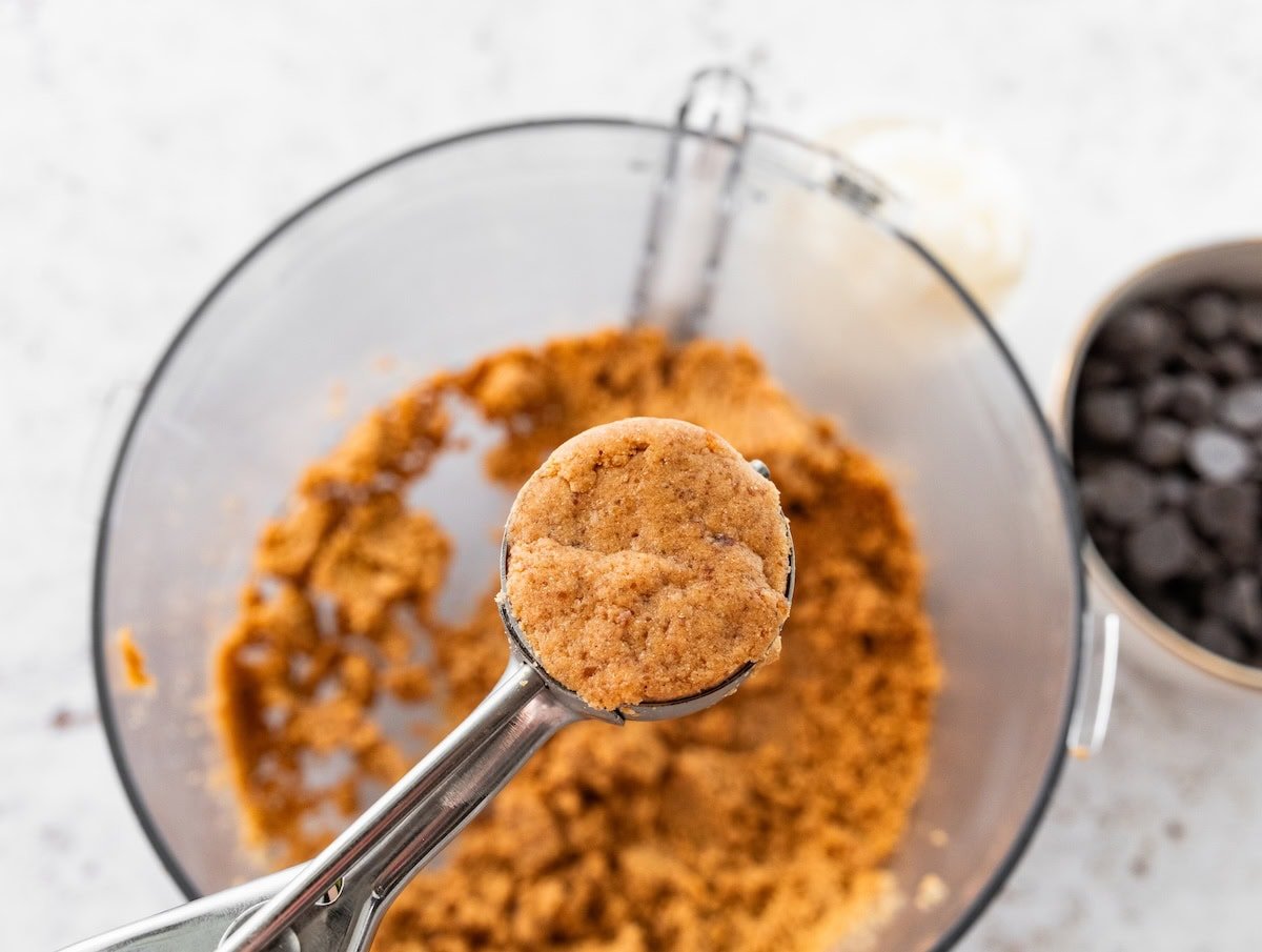 A small cookie scoop holding peanut butter buckeye dough over a food processor bowl.