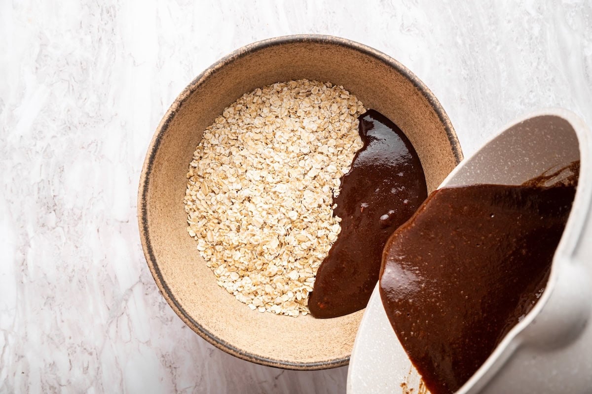 Melted chocolate being poured from a large bowl into another bowl that is full of quick oats.