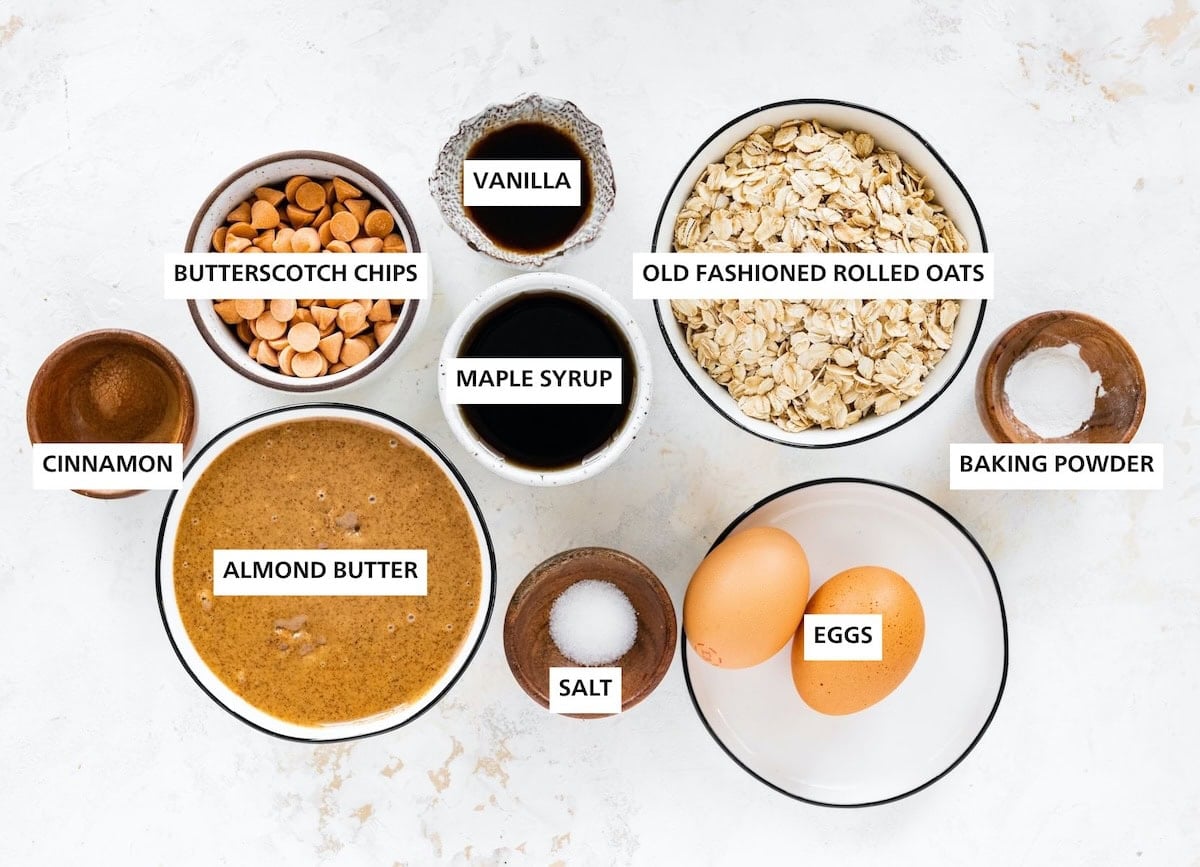 Ingredients measured out to make Oatmeal Scotchies: baking powder, oats, eggs, vanilla, maple syrup, salt, butterscotch chips, almond butter and cinnamon.