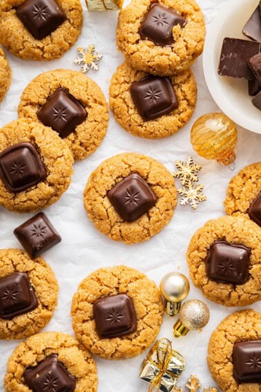 Peanut butter blossom cookies arranged on white parchment with small gold ornaments, and extra chocolate pieces scattered around.
