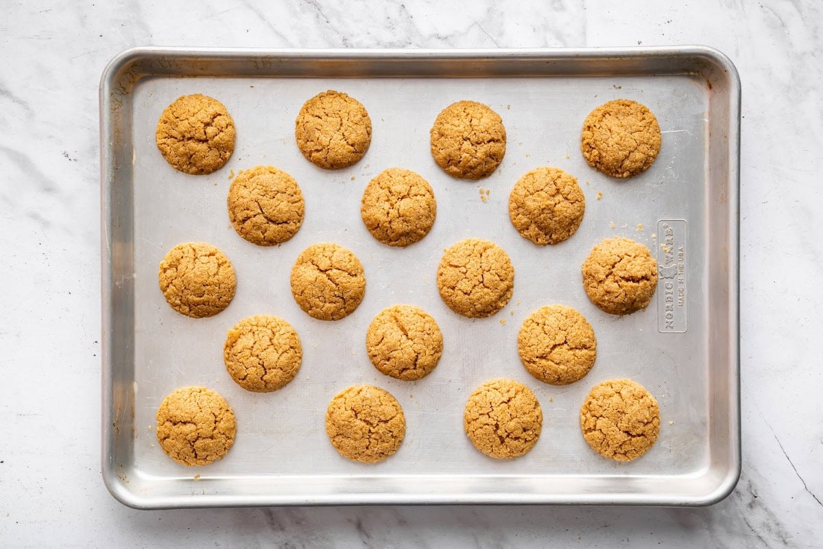 Freshly baked peanut butter cookies resting on a metal baking sheet, each one lightly cracked on top and waiting for the chocolate to be added.