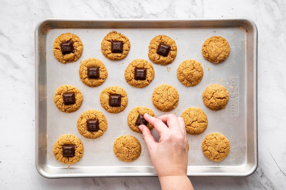 A hand gently pressing a square of chocolate into the warm center of a peanut butter cookie on a baking sheet, while several cookies around it already have their chocolate pieces added.