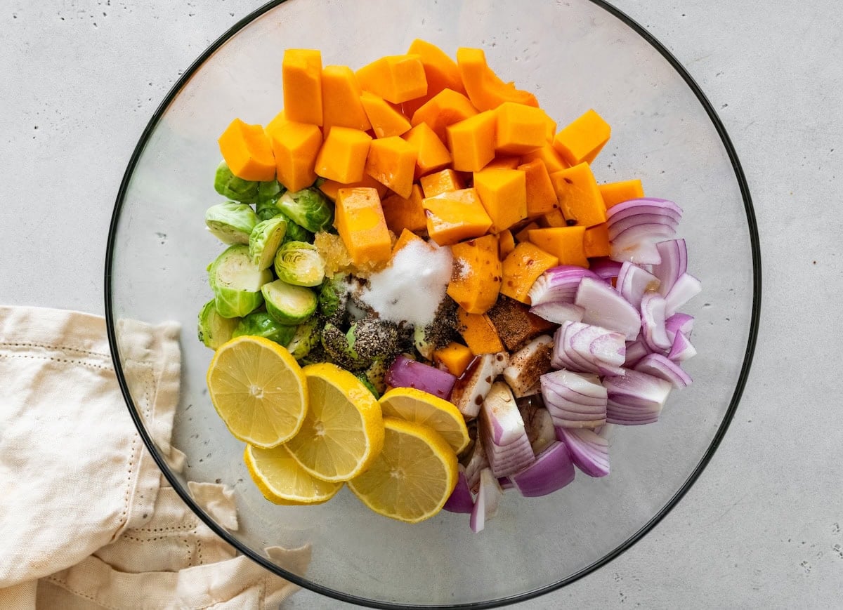 Chopped ingredients Chopped butternut squash, red onion, brussels sprouts and lemon in a glass mixing bowl sprinkled with seasonings and oil.