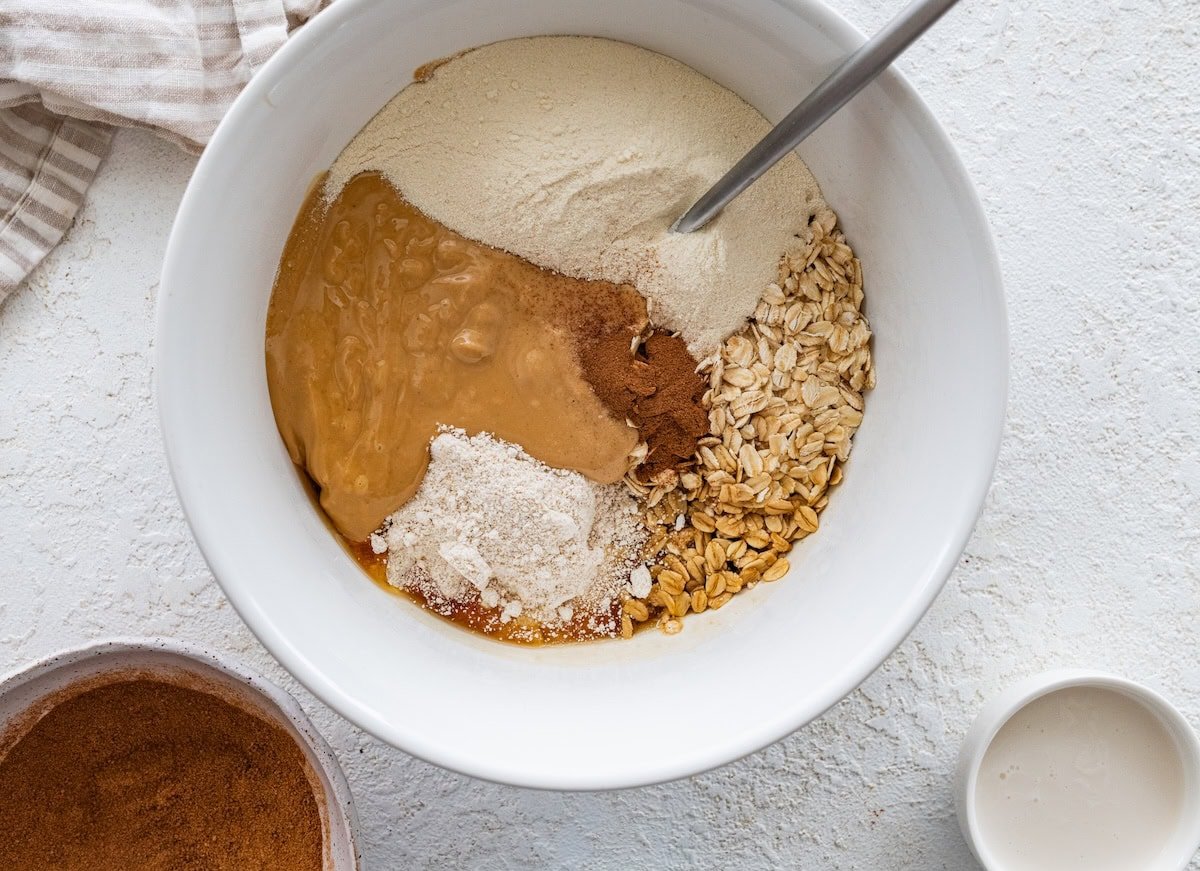 Mixing bowl filled with oats, oat flour, cashew butter, protein powder and cinnamon before stirring.
